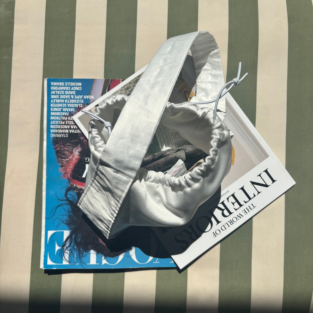 A small silver satin bucket bag with a top handle, resting on top of two magazines on a green and cream striped cushion.
