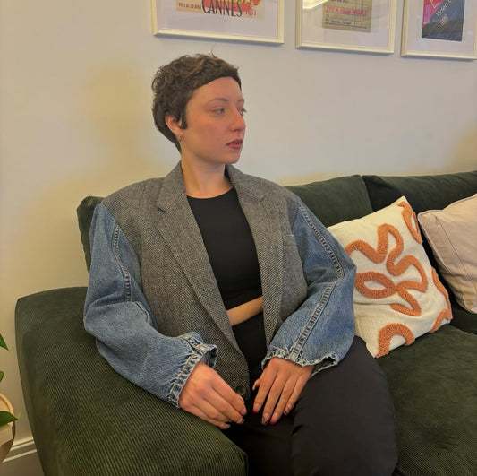 Person sitting on a couch wearing a gray blazer with denim sleeves, in a room with framed pictures on the wall.