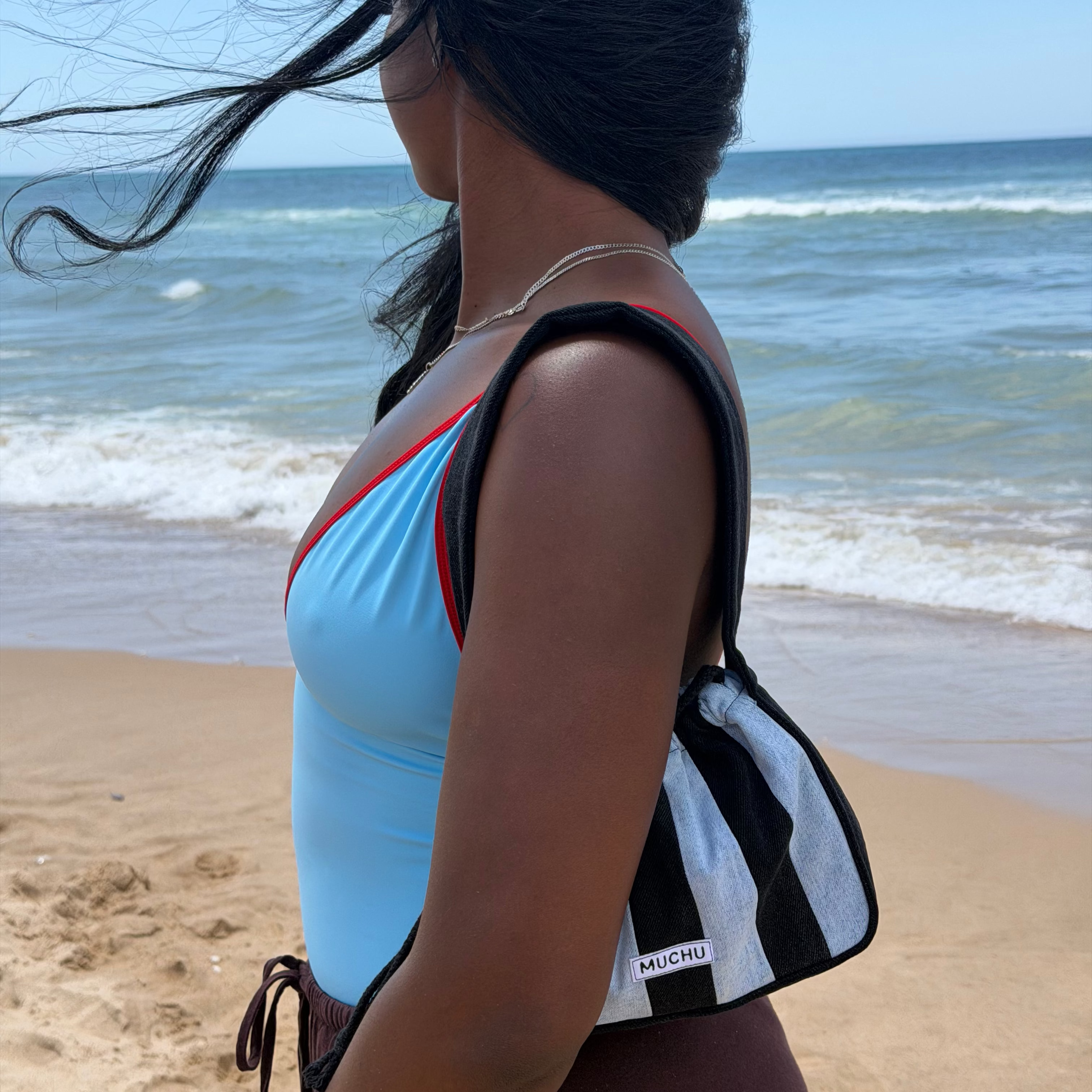 A woman on a sandy beach with the ocean in the background, wearing a light blue swimsuit and carrying a black and white striped shoulder bag.